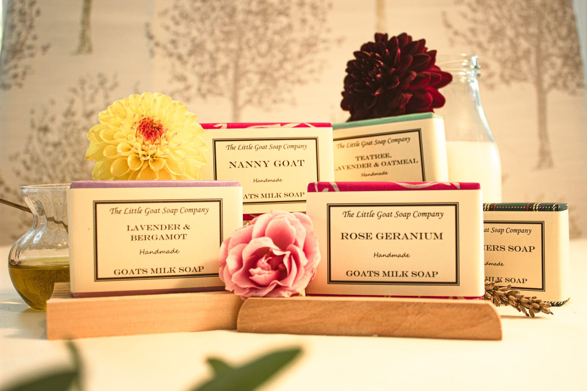 large range of soaps from the little goat soap company, set dressed with a bottle of goats milk, yellow and red flowers. The soaps are arranged on soap racks.