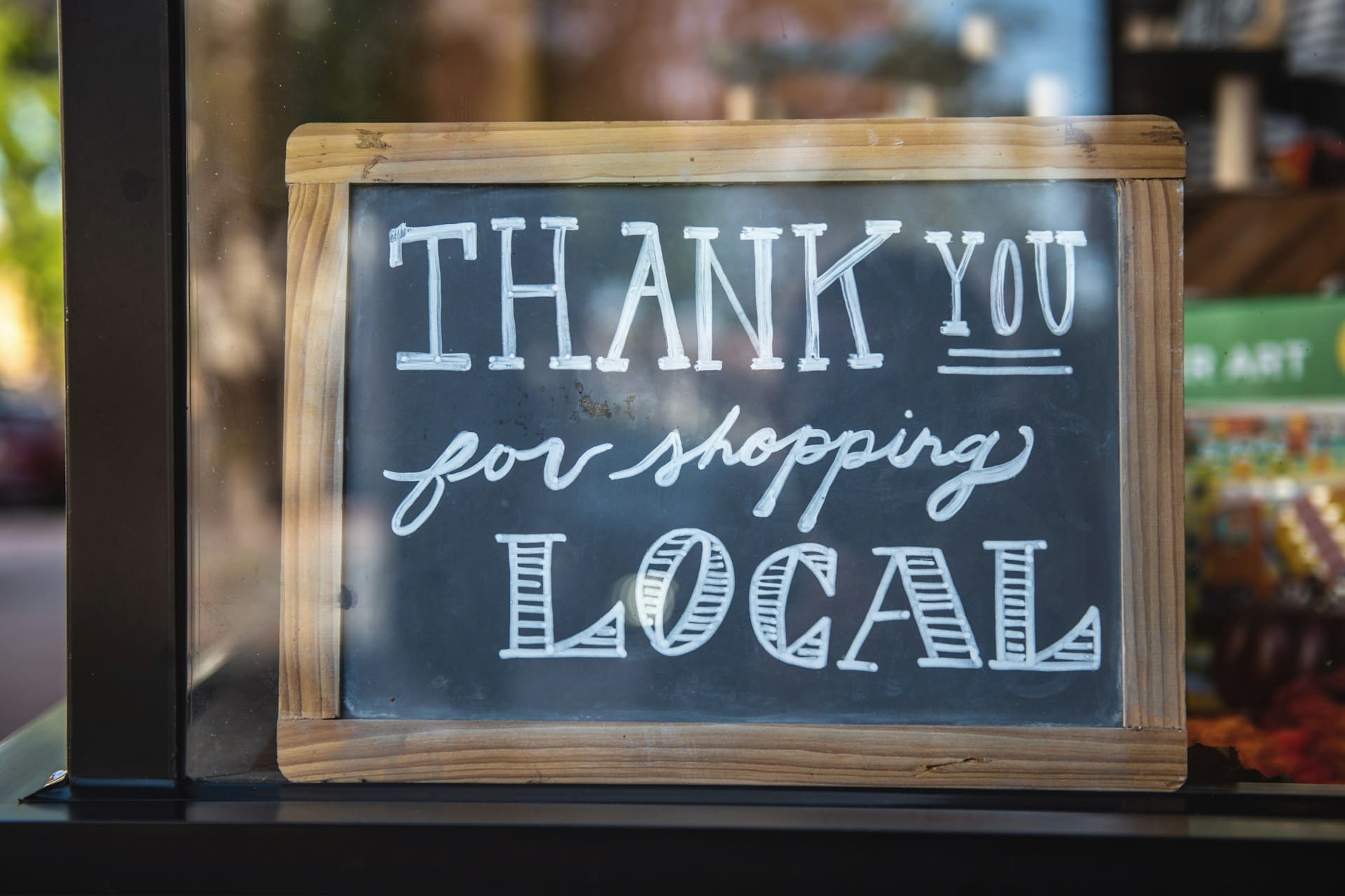A wooden chalkboard in a shop window with the words 'thank you for shopping local' written in white chalk